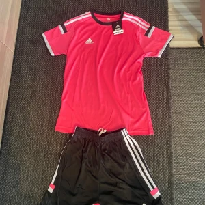 Rosa Adidas träningströja med svarta detaljer och matchande shorts - Säljer en rosa  Adidas fotbollströja med svarta och vita detaljer på axlarna. Tröjan är kortärmad och tillverkad i lätt och ventilerande Climacool-material. Ingår med matchande shorts! Perfekt för träning eller match. Klassisk Adidas-logga på bröstet och på shortsen. För ENDAST 100kr så får du de bästa träningskläder! Pris kan diskuteras.
