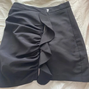 Svart kjol med volang och dragkedja - Säljer en svart kortkjol med snygg volangdetalj och dragkedja framtill. Kjolen har en stilren design med en cool twist tack vare volangen som ger extra attityd. Perfekt för dig som gillar trendiga plagg med unika detaljer.