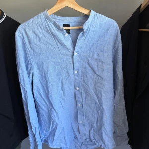 Ljusblå skjorta med ståkrage från H&M - Säljer en ljusblå skjorta från H&M med ståkrage och knappar framtill. Skjortan har en bröstficka och är tillverkad i ett lätt bomullstyg. Perfekt för en avslappnad men stilren look.