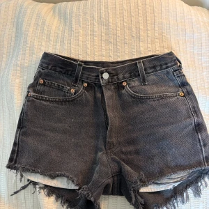 Svarta jeansshorts från Levi's - Säljer ett par svarta jeansshorts från Levi's med fransig kant och klassiska fem fickor. Shortsen har hög midja och knappgylf. Perfekta till sommaren för en avslappnad look.