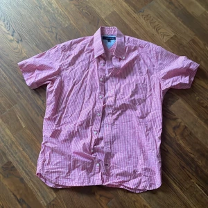 Rosa och vit rutig kortärmad skjorta från Tommy Hilfiger - Kortärmad skjorta från Tommy Hilfiger i rosa och vitt rutigt mönster. Skjortan har klassisk krage, knappar framtill och en liten broderad logga på bröstfickan. Insidan av knappslån har ett färgglatt mönster. Den kanske ser lite skrynklig ut men det blir bättre om man bara stryker