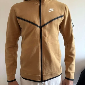 Beige brun zip-hoodie från Nike - Säljer en beige brun kofta från Nike med svart dragkedja och svarta detaljer över bröstet. Jackan har huva, lång ärm och Nike-logga på bröstet. Perfekt för en sportig och avslappnad stil.