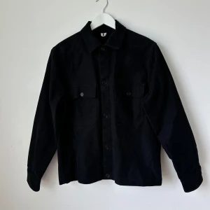 Svart skjortjacka från Arket - Stilren svart skjortjacka från Arket med klassisk krage och knäppning framtill. Två bröstfickor med knappar ger en clean look. Perfekt att bära som lager över t-shirt eller hoodie. Artnr: Ö291