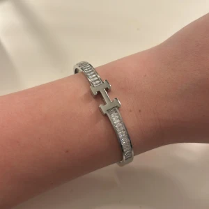 Silverfärgat armband med H-detalj - Stilrent armband i silverfärg med en tydlig H-formad detalj framtill. Armbandet har en glittrig yta längs bandet som ger extra elegans. 