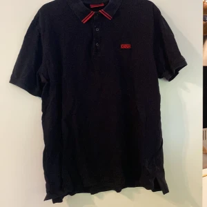 Svart pikétröja från Hugo Boss - Säljer en svart pikétröja från Hugo Boss med röda detaljer på kragen och röd broderad logga på bröstet. Tröjan har klassisk krage med knappar och är kortärmad. Perfekt för en stilren look.