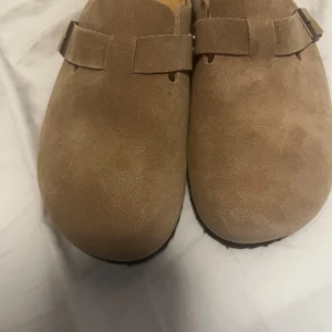 Beige mocka Birkenstock tofflor - Säljer ett par klassiska Birkenstock tofflor i beige mocka med svart mönstrad sula. Den är helt nya och oanvända. Råka slänga kartongen så kan inte lämna den tillbaka, de är i storlek 44 