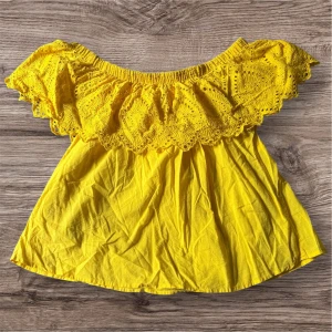Gul offshoulder blus med spets från Gina Tricot - Säljer en solgul offshoulder blus från Gina Tricot med vacker spetsvolang upptill. Blusen har en luftig passform och är perfekt för varma dagar. Supersnygg att styla till både jeans och kjol!