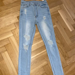 Ljusblå slitna skinny jeans - Säljer ett par ljusblå skinny jeans med slitningar på framsidan. Jeansen har hög midja och dragkedja med knapp. Perfekta för en avslappnad och trendig look.