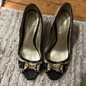 Svarta pumps med guldiga detaljer från Guess - Säljer ett par eleganta pumps från Guess med svart lackad tå, mönstrad canvas med Guess-logga och snygga guldiga kedjedetaljer framtill. Skorna har klack och rund tå, samt dekorativ rosett och ringdetalj.