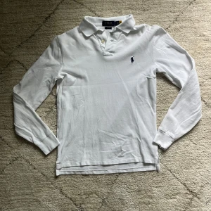 Vit långärmad pikétröja från Polo Ralph Lauren - Klassisk vit långärmad pikétröja från Polo Ralph Lauren i slim fit-modell. Tröjan har krage, knappar vid halsen och den ikoniska broderade loggan på bröstet. andvänd fåtal gånger med inga defekter!!!! 