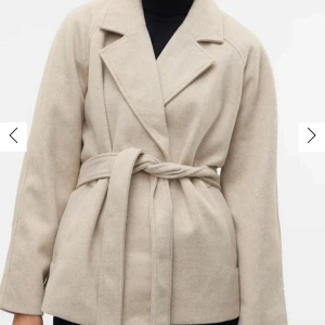 Beige kappa med bälte - Stilren beige kappa med knytbälte i midjan och klassisk krage. Jackan har lång ärm och en rak, något oversized passform. Perfekt för lager-på-lager och enkel att matcha med olika outfits. Från Veromoda!