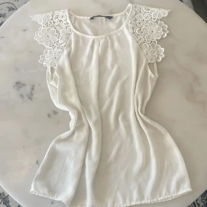 Vit blus med spetsärmar från Zara Basic - Säljer en vit blus från Zara Basic med vackra spetsdetaljer på axlarna. Blusen är ärmlös och har en rund halsringning. Perfekt för en fräsch och stilren look.