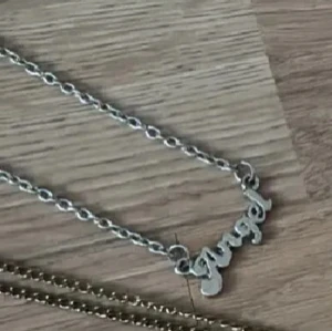 Silverfärgat halsband med texten 'angel' - Säljer ett trendigt halsband i silverfärg med kedja och ett hängsmycke där det står 'angel' i skrivstil. Perfekt accessoar för att lyfta din outfit med en personlig touch.