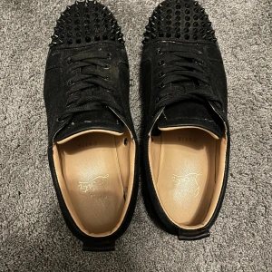 Svarta sneakers med nitar och röd sula - Svarta sneakers med mockaliknande material, snörning och rund tå. Skorna har svarta nitar på tån och en ikonisk röd sula undertill. Perfekta för dig som vill sticka ut med en edgy stil.