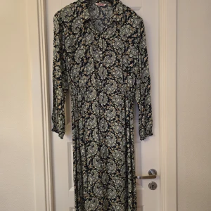 Mönstrad långklänning från Indiska - Säljer en långklänning från Indiska med blommigt mönster i blått, beige och grönt. Klänningen har lång ärm med resår vid ärmslut, krage och knappar framtill. Perfekt för dig som gillar bohemisk stil.