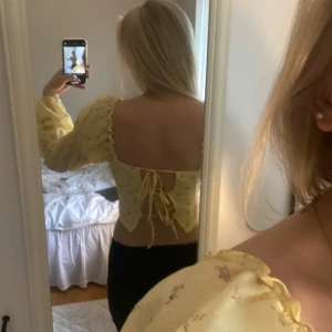 Gul blommig croppad topp med puffärmar - Söt gul croppad topp med små blommor och stora puffärmar. Toppen har öppen rygg med snörning och volangkant vid ärmslut och nederkant. Perfekt för vår och sommar!