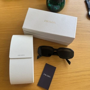Svarta solglasögon från Prada - Säljer ett par svarta, rektangulära solglasögon från Prada. Glasögonen har breda skalmar med logotypen i vitt på sidan. Kommer med originalfodral och ask i vitt. Snygg och stilren accessoar nypris ca 3000kr