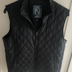 Svart quiltad väst från Nimbus - Stilren svart quiltad väst från Nimbus med dragkedja framtill och två fickor. Perfekt för lager-på-lager och passar till många olika outfits. Klassisk design med hög krage och diskret mönster.