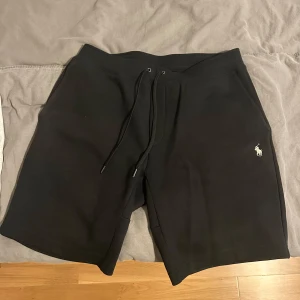 Svarta shorts från Polo Ralph Lauren - Svarta shorts från Polo Ralph Lauren med klassisk broderad logga på ena benet. De har snörning i midjan och en bakficka. Perfekta för en avslappnad stil och tillverkade i mjukt material.