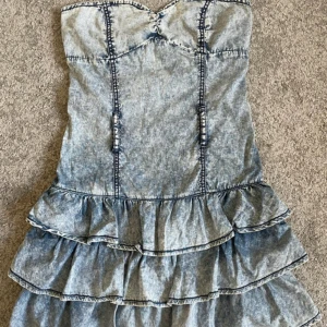 Jeansklänning med volanger - Säljer en snygg jeansklänning med volanger och smock i ryggen. Klänningen är ärmlös och har en figurnära passform