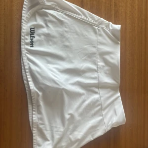 Vit tenniskjol från Wilson - Säljer en klassisk vit tenniskjol från Wilson i storlek M. Kjolen har en sportig design med diskret Wilson-logga framtill och är tillverkad i ett lätt och stretchigt material som passar perfekt för aktiva dagar på tennisbanan.