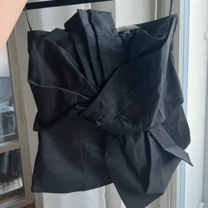 Svart asymmetrisk tubtopp med rosett - Säljer en svart tubtopp med en rosett framtill. Toppen har en dragkedja i ryggen och är använd en kväll! 🩷