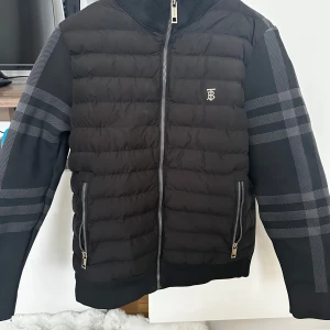 Svart pufferjacka från Burberry - Säljer en svart pufferjacka från Burberry med klassiskt rutmönster på ärmar och insida. Jackan har dragkedja framtill, två fickor med dragkedja och ribbade muddar. Perfekt för kyliga dagar och snygg till streetwear.