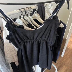 Svart croppad linnetopp med volang - Svart linnetopp med smala axelband och volangdetaljer längs urringningen. Toppen är croppad och har en luftig passform, perfekt för varma dagar.