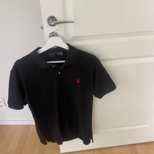 Svart pikétröja från Polo Ralph Lauren - Hej säljer nu min svart pikétröja från Polo Ralph Lauren med röd broderad logga på bröstet. Tröjan har bara används några få gånger och har inga tecken på användning eller defekter.