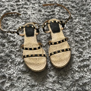 Beiga sandaler med nitar och svarta remmar - Säljer ett par beiga sandaler från H&M med svarta remmar dekorerade med guldfärgade nitar. Skorna har platt sula i flätat material och flera tunna remmar som går runt foten och ankeln.