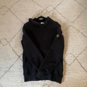 Svart hoodie från Stone Island  - Säljer en svart hoodie från Stone Island Junior med klassisk logga på ärmen. Tröjan har huva och magficka, perfekt för en avslappnad stil. Mjuk insida och ribbade muddar vid ärmslut och nederkant.