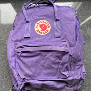 Lila ryggsäck från Fjällräven Kånken - Säljer en klassisk lila Fjällräven Kånken ryggsäck med det ikoniska runda märket framtill. Väskan har ett stort huvudfack, en framficka med dragkedja och två sidofickor. Praktiska handtag upptill och justerbara axelremmar.