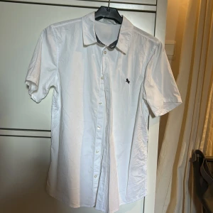 Vit kortärmad skjorta med broderad detalj - Säljer en klassisk vit skjorta med korta ärmar och knappar framtill. Skjortan har en liten broderad logga på bröstet och en stilren krage. Perfekt för en fräsch och enkel look. Den är använd en gång.