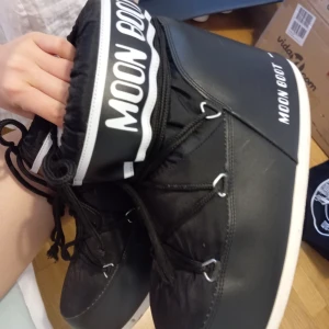 Svarta Moon Boots - Säljer ett par klassiska svarta Moon Boots med vit sula och vit logga. Skorna har snörning framtill och är fodrade på insidan för extra värme. Perfekta för kalla vinterdagar och snöiga äventyr. Storlek 43-45