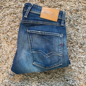 Replay Anbass jeans - Säljer ett par blå Replay Anbass jeans med klassisk femficksdesign och snygga slitningar. Jeansen har normal passform och raka ben, med Replay-logga på bakfickan och läderpatch i midjan. Perfekta för dig som gillar stilrena och bekväma jeans.