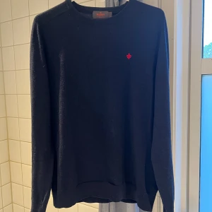 Navy  merinoullströja från Morris - Säljer en klassisk navy tröja från Morris i 100% merinoull. Tröjan har rund halsringning, lång ärm och en liten röd broderad logga på bröstet. Perfekt för en stilren och enkel look.
