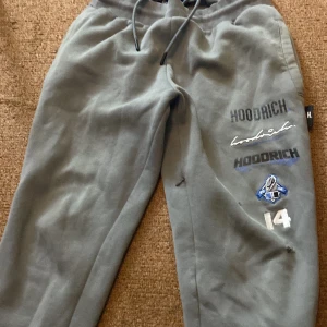 Grå mjukisbyxor från Hoodrich - Snygga grå mjukisbyxor från Hoodrich med tryck och broderade detaljer på benen. Byxorna har resår i midjan med snörning och en bakficka med Hood Series-logga. Perfekta för en avslappnad stil. Med två små hål i vänstra sida ben