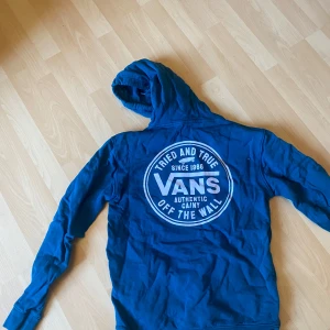 Blå hoodie från Vans - Säljer en blå hoodie från Vans med stor vit logga på ryggen och mindre logga på bröstet. Tröjan har huva och en stor magficka framtill. Perfekt för dig som gillar streetwear och vill ha något bekvämt och snyggt.