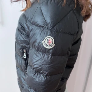 Svart tunn dunjacka från Moncler - Säljer en tunn svart dunjacka från Moncler med hög krage och två dragkedjeförsedda fickor framtill. Jackan har quiltad design och Moncler-logga på ärmen. Perfekt för kyliga dagar och riktigt snygg till streetwear. Finns tre små hål som lätt går att ordna med textiltejp. Storlek 152 men tycker den passar 140-156