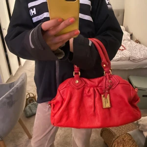 Röd handväska från Marc by Marc Jacobs - Säljer en snygg röd handväska från Marc by Marc Jacobs med guldfärgade metalldetaljer och logga framtill. Väskan har både handtag och axelrem samt ett stort fack med dragkedja och mönstrat innerfoder med text. Perfekt för dig som vill sticka ut! Knappt använd