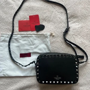 Valentino camera bag - Säljer en svart axelväska från Valentino Garavani med silverfärgade och lite svarta nitar runt kanterna. Väskan har justerbar axelrem och logga framtill. Inga defekter och knappt använd, äktehetsbevis med.
