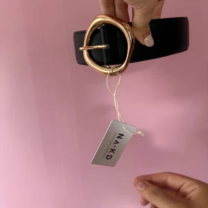 Svart bälte med guldspänne från NA-KD - Stilrent svart bälte från NA-KD med ett unikt, rundat guldspänne. Perfekt accessoar för att lyfta din outfit. Bältet har en bred design och klassisk look.