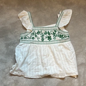 Vit blus med gröna broderier och volangärmar - Säljer en söt vit blus med gröna broderier och volangärmar. Blusen  har en knytning i ryggen och ett vackert blommönster framtill. Perfekt för varma dagar! Hör av er vid frågor❤️