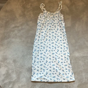 Vit blommig sommarklänning med axelband - Söt vit klänning med små blå blommor och twistade axelband. Lätt och luftig modell som passar perfekt till varma dagar. Klänningen har en rak passform och är ärmlös, med en längd som går till knäna. Den har en enstaka liten fläck men de är inte så synliga! Hör av er vid frågor❤️