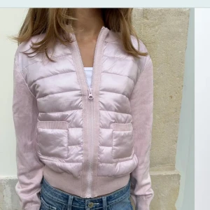 Ljusrosa pufferjacka med stickade ärmar - Säljer en ljusrosa pufferjacka med stickade ärmar och ribbade muddar. Jackan har dragkedja framtill och två fickor. Perfekt för lager-på-lager och en snygg detalj till jeans.