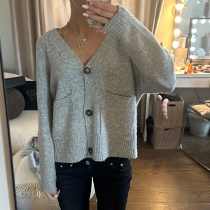 Grå stickad kofta med knappar - Mysig grå stickad kofta med stora knappar framtill och två fickor. Koftan har en avslappnad passform och ribbade muddar vid ärmslut och nederkant. Perfekt att slänga på sig över en t-shirt eller linne.