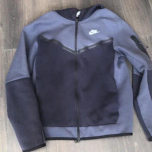 Nike tech - Säljer en mörkblå zip-jacka från Nike med svart V-detalj över bröstet och liten logga på vänster sida. Jackan har lång ärm och dragkedja framtill, samt en ficka med dragkedja på ena ärmen. Perfekt för en sportig look.