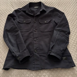 Mörkblå overshirt från Gabba - Säljer en stilren mörkblå overshirt från Gabba med klassisk krage och två bröstfickor med knappar. Skjortan har lång ärm och knäppning framtill, perfekt att bära som lager över t-shirt eller tröja.