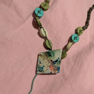 Färgglatt halsband med knappar och motivberlock - Unikt halsband med stora runda knappar i grönt, turkost och vitt. Halsbandet har en fyrkantig berlock med färgglatt motiv. Knapparna är trädda på grå snodd och halsbandet har justerbar längd.