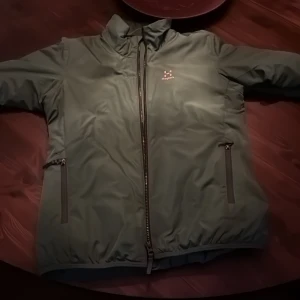 Grön vindjacka från Haglöfs - Säljer en grön vindjacka från Haglöfs med dragkedja framtill och två fickor med dragkedja. Jackan har hög krage och enkel, stilren design. Perfekt för utomhusaktiviteter och vardagsbruk. Pris kan diskuteras!